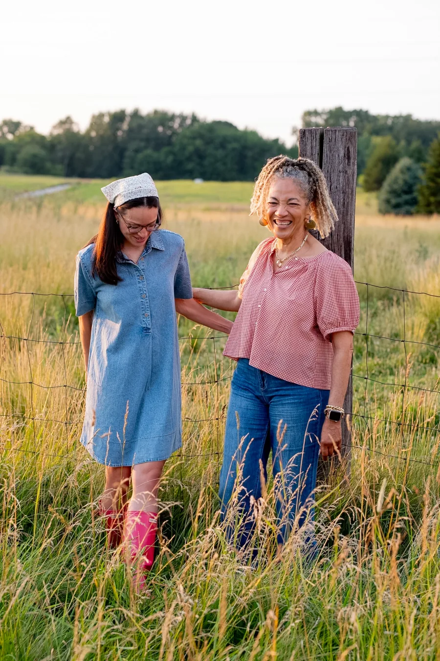 Two women laughing together beside a fence in a sunlit field